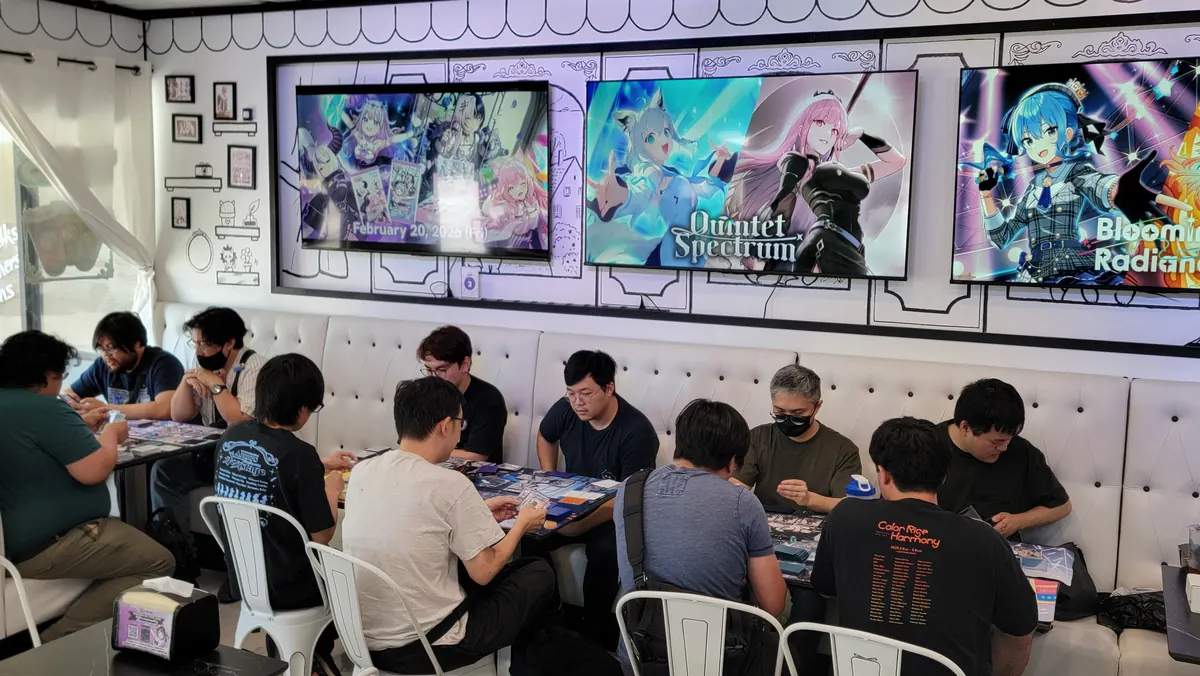Players at a hololive OCG tournament in the café dining room, with hololive art on the TVs above the booth seats