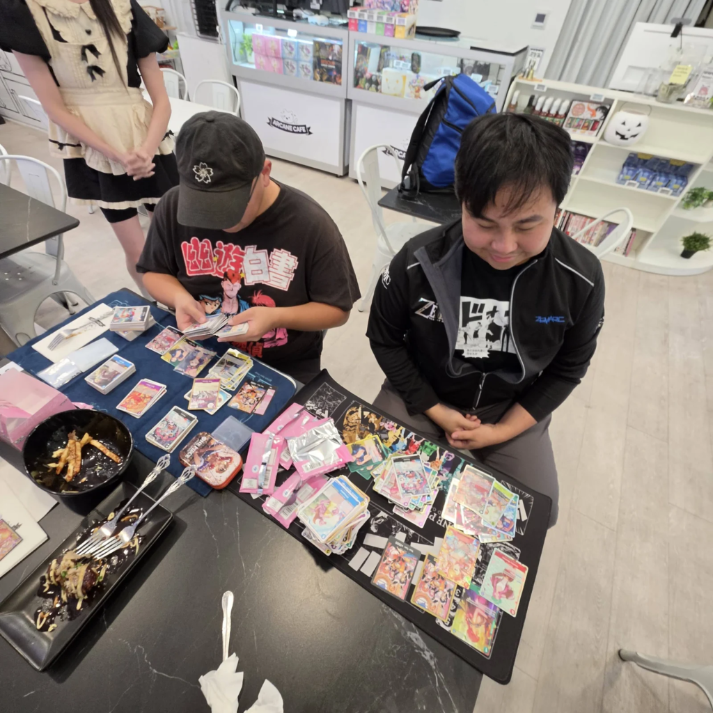 Guests opening One Piece Card Game EB-05 Heroines Edition vol.2 booster packs at a table with food while a maid stands nearby, EB-05 launch and pull day at Arcane Maid Cafe, Arcadia CA